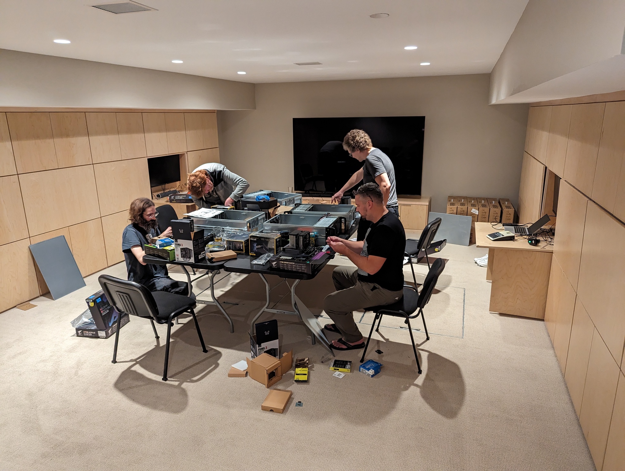 A photo depicts the game room from earlier. Four guys are standing around two folding tables set up in the middle of the room, where they are assembling the computers. Each person has an open chassis in front of him with components and boxes strewn about.