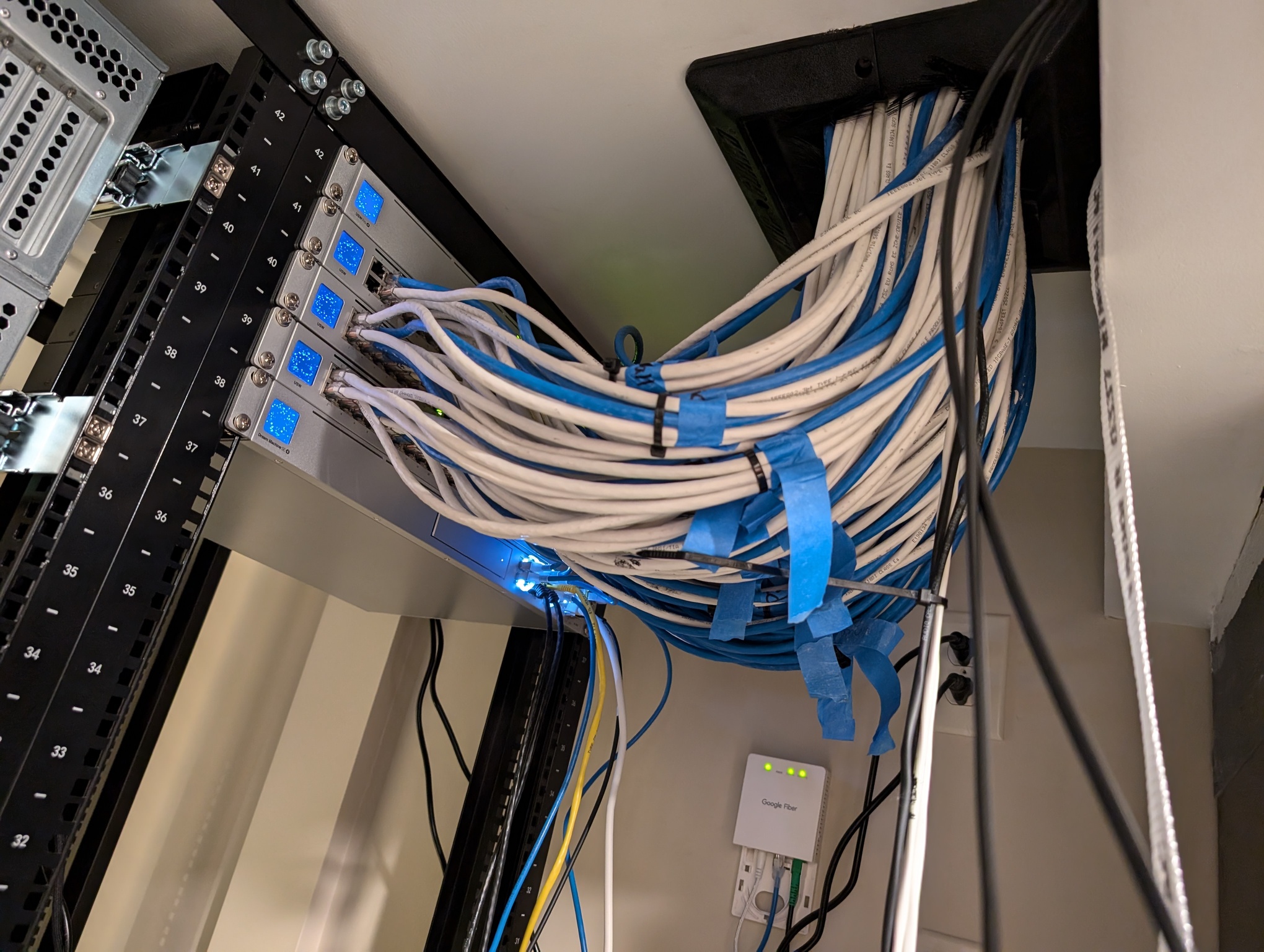 A photo shows an up-close view of a stack of five network switches mounted at the top of the rack furthest from the door. An enormous bundle of category 6 network cables comes out of a hole in the ceiling and connects to the network switches. A Google Fiber box is mounted on the wall nearby.