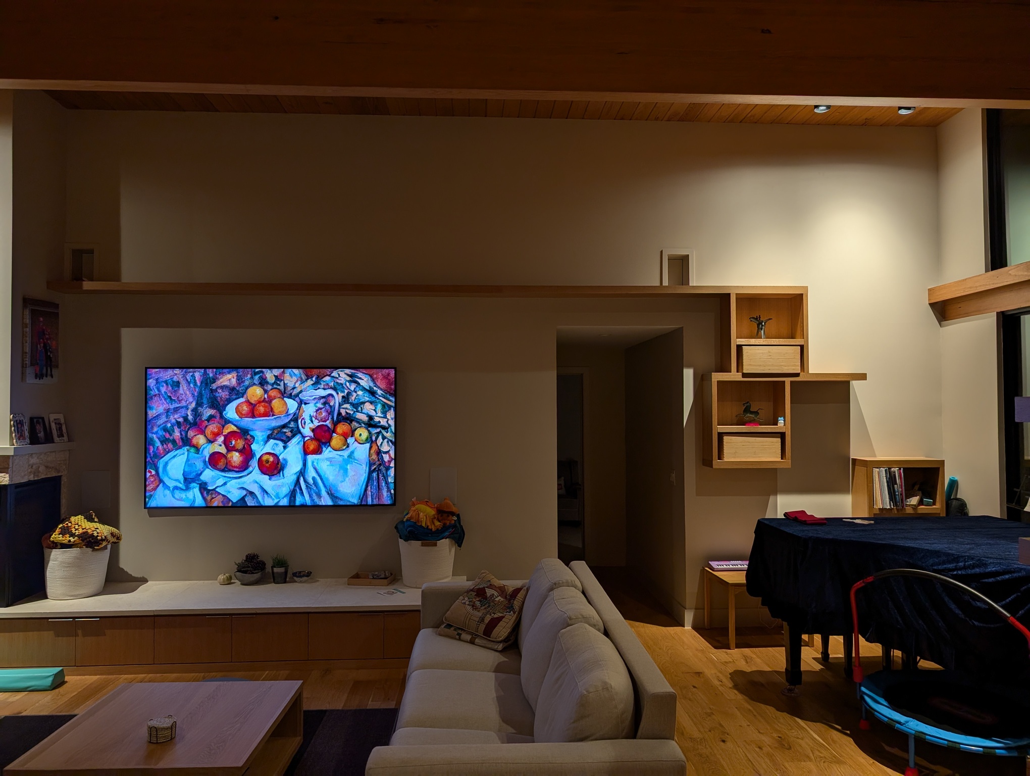 A photo depicts the living room again, showing a wall that wasn't visible before. The wall features at 85 inch TV displaying artwork. To the right of the TV is a hallway entrance. Above both of these, a long wood shelf extends across the length of the wall. To the right of the hallway entrace is more wall space, where more wood shelves are arranged such that a cat could hop up from shelf to shelf to get to the top. At either end of the long top shelf are two cat doors through the wall.