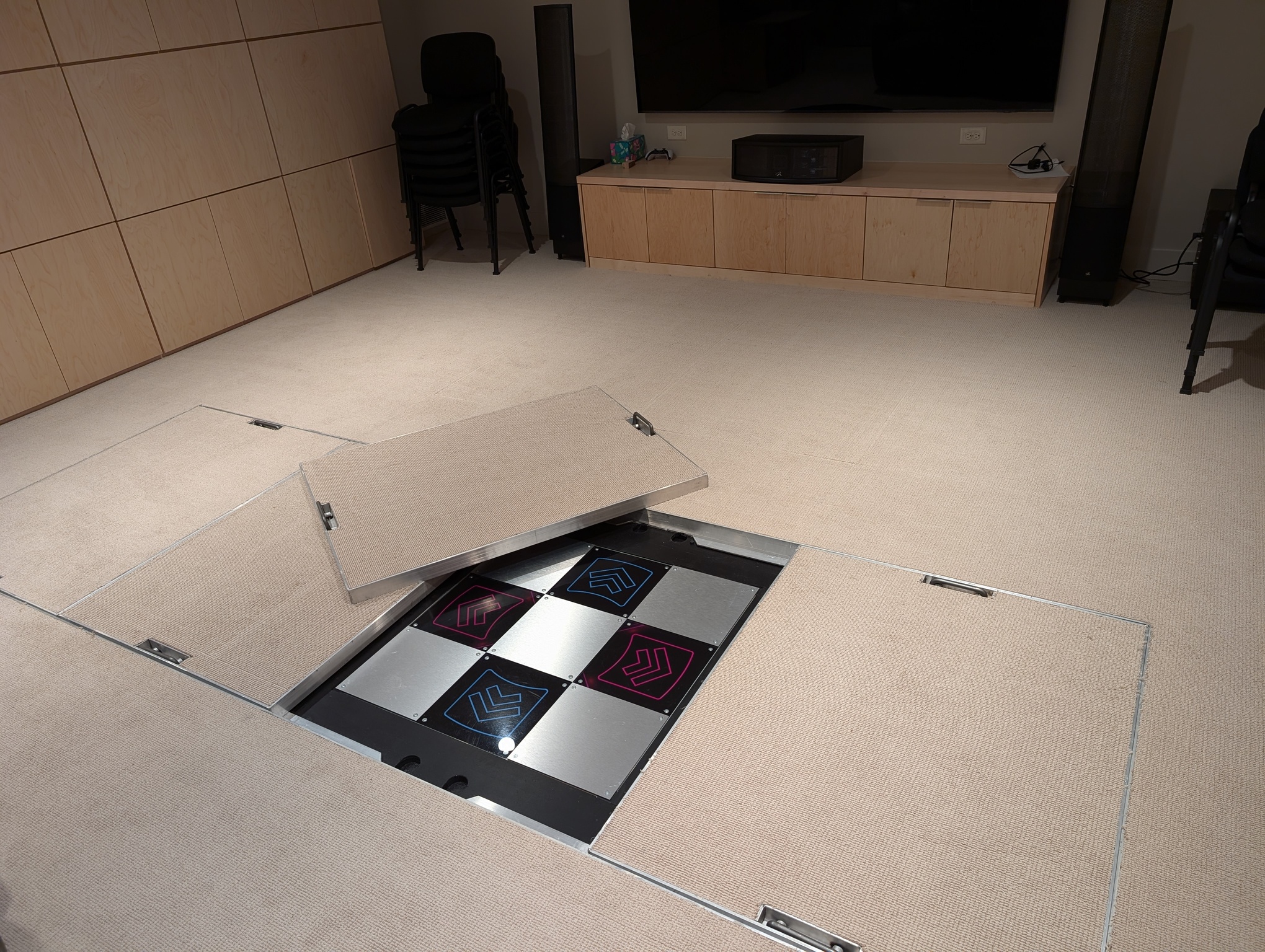 A photo shows the floor of the game room. The floor has a row of four removable panels. The panels are aluminum with carpeting on top matching the rest of the floor and retractable handles at either end. One of the panels has been lifed and placed to the side, revealing a Dance Dance Revolution game pad underneath.