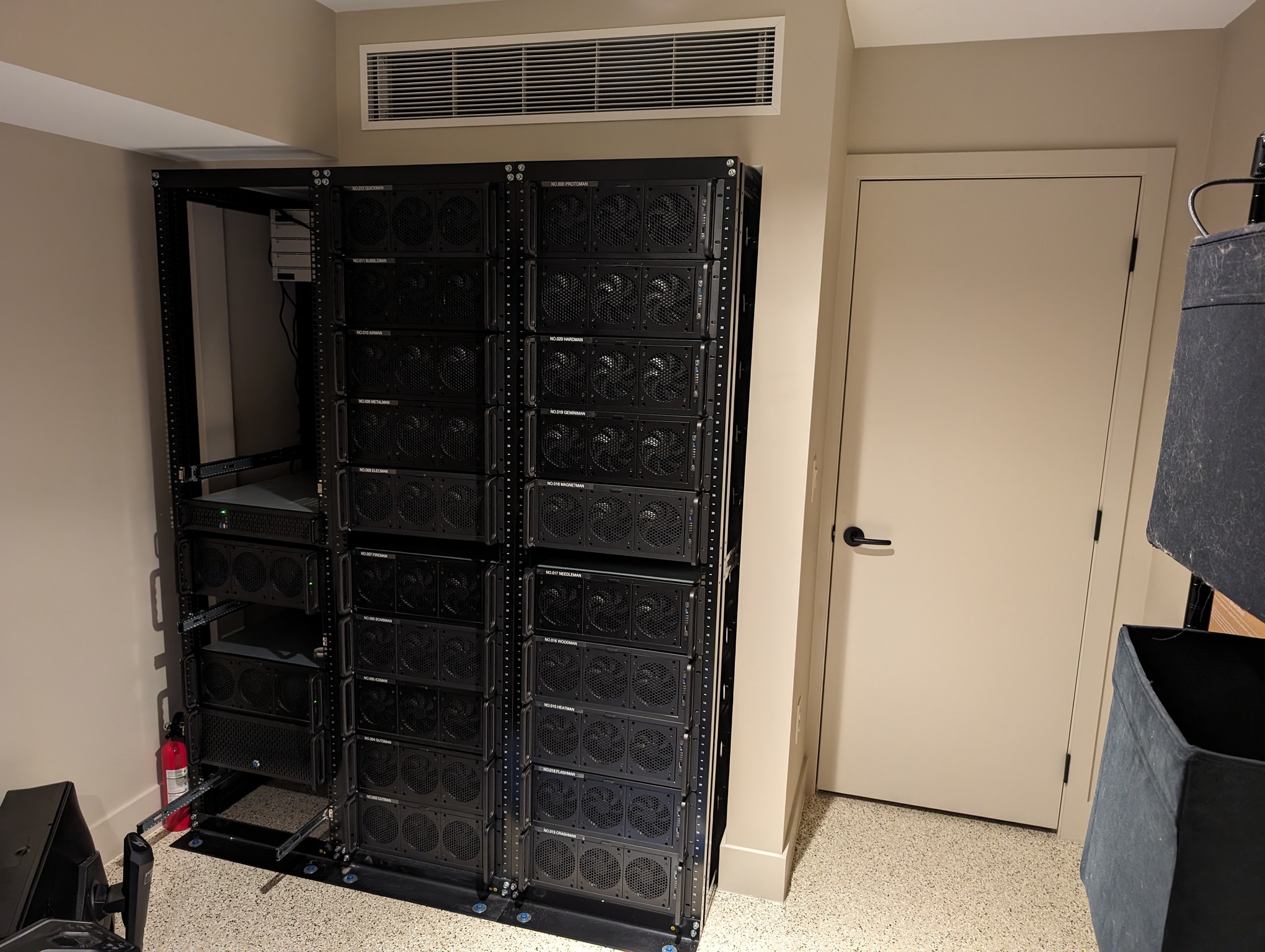 The photo depicts a room with three computer racks side-by-side. The center and right racks each hold ten computers; these are the game machines hooked up to the monitors seen in previous photos. The right rack is mostly unpopulated, but contains two computers that are our personal desktops, and a server machine, and one chassis that is currently empty. All of the machines except the server are in 4U rackmount chasis; the server is 2U. The racks are embedded in the wall. To their right is a closed door which leads to their back side. Above the racks is an air vent which provides cold air from a dedicated air conditioner; the intake is on the other side of the machines.