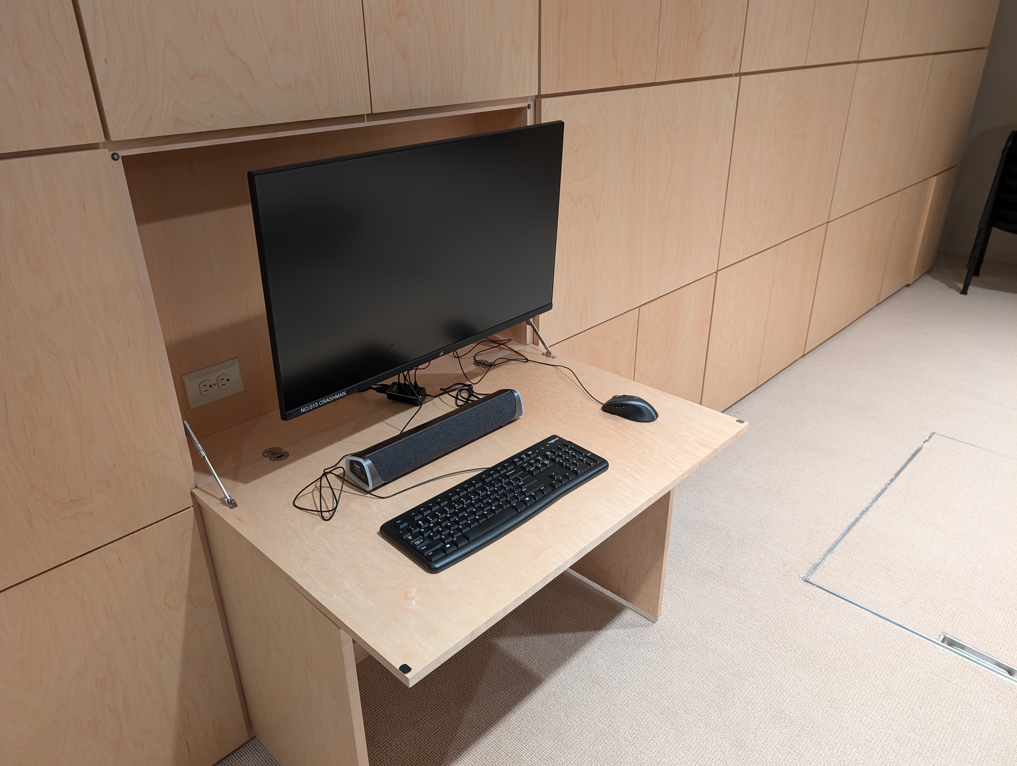 A photo shows an up-close view of the game stations that lined the left wall in the previous photo. In this photo, one station is open, while the others are all closed. The closed stations merely look like wood paneling on the wall; the monitors are entirely hidden. The open station shows that a panel in front of the monitor folds outwards and down to become the desk on which the keyboard sits. Two panels below this one swing outwards to form supports for the left and right sides of the desk. The monitor is on a retractable mount such that it can be pulled away from the wall, closer to the user. There is a power outlet behind the monitor, suitable for charging a phone while sitting at the station.