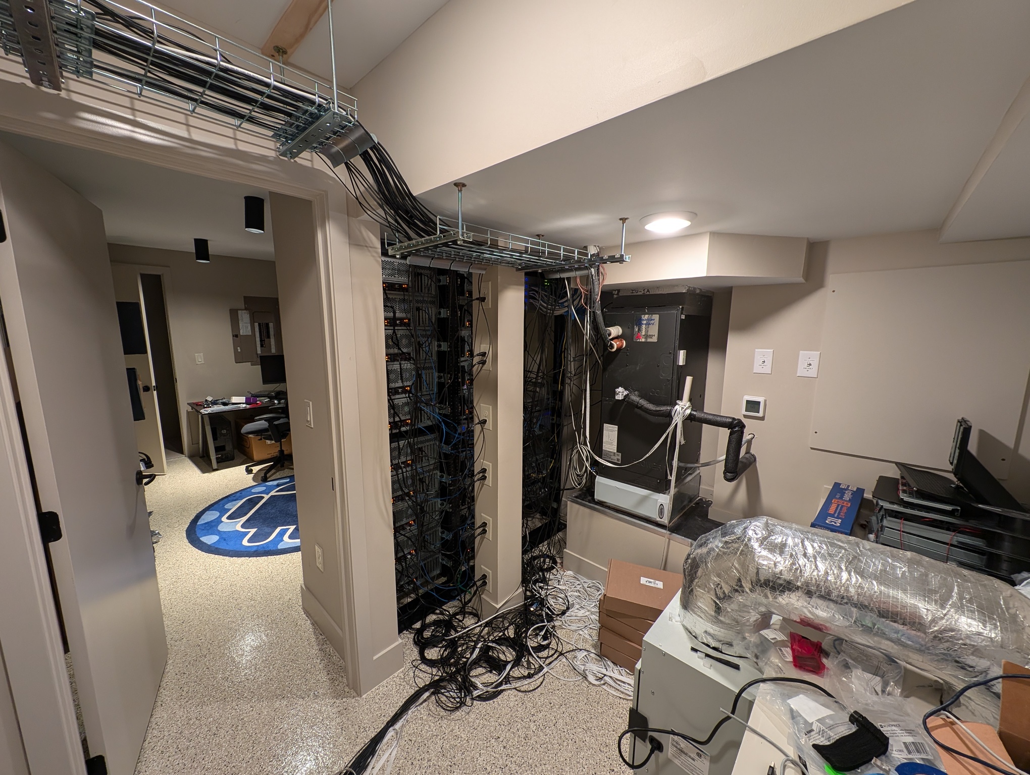 A photo depicts the back side of the computer racks. To their left is the door, now open, leading to the front side. Many wires are visible in the photo, some organized in a cable track hanging from the ceiling, others strewn haphazardly on the floor. Directly behind the server racks there is a column full of power outlets which many of the machines are plugged into. Also visible in the picture is various HVAC equipment, inculding an air handler and a HEPA filtration unit. These actually don't serve this room but were placed here because they are ugly.