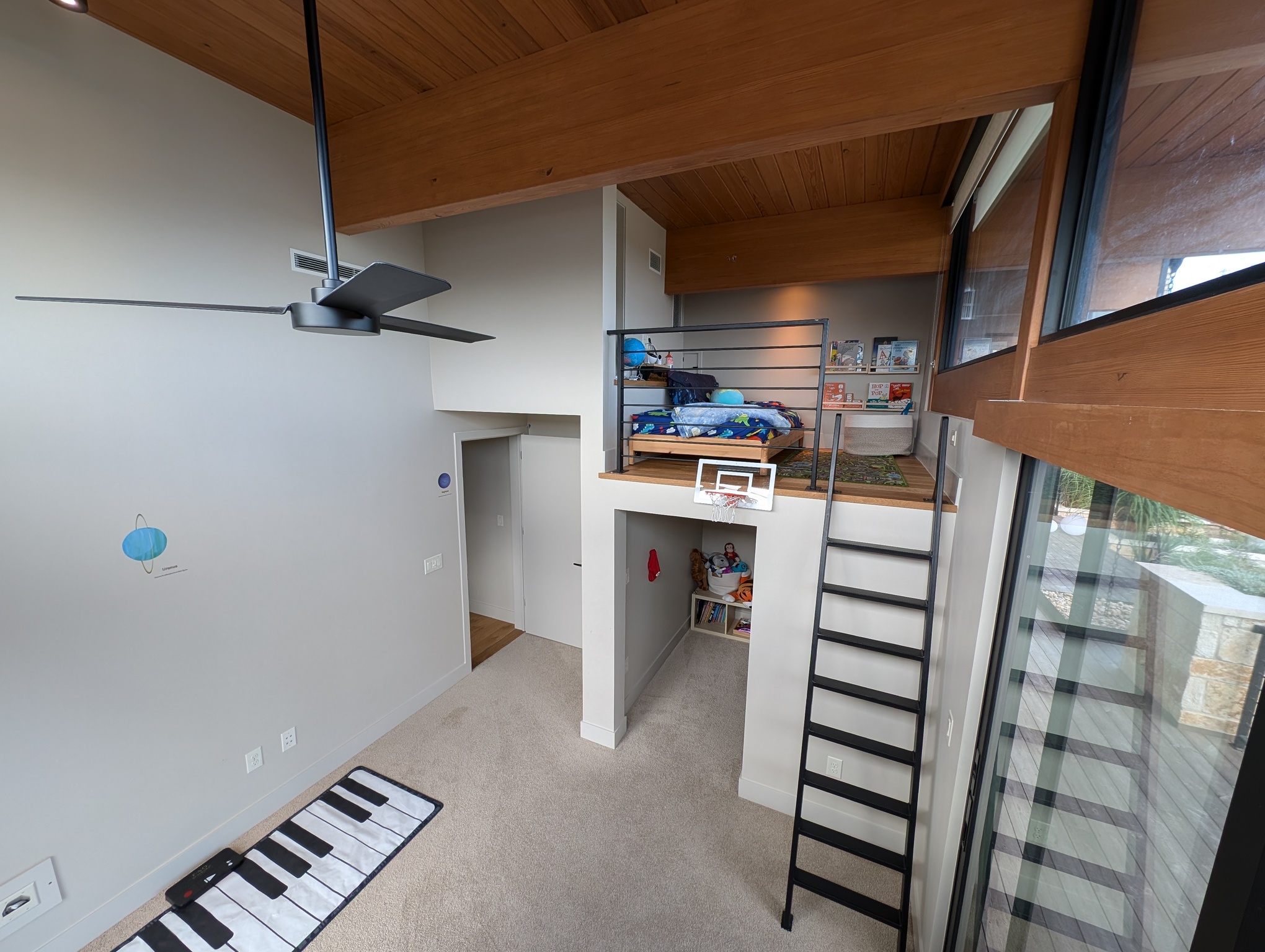 A photo depicts a kid's bedroom. The room features 14-foot ceilings similar to the living room. The right side of the room has two levels, a 6-foot-tall walk-in closet and a loft above it. A black metal ladder provides access to the loft. The loft is about the size of a queen mattress, but currently features a twin bed. To the right of the bed is a small bookshelf with children's books. To the left of the bed is a narrow ledge along the wall. At the back of this ledge is the other side one of the cat doors leading to the living room. Also above the ledge, a door leads to a kid-sized hallway which leads to the other kid's loft. On the opposite side of the room from the loft, a wall box provides access to a cable conduit leading to the engine room, but nothing is currently connected through it. A computer desk could be placed here when the child is old enough to have one.