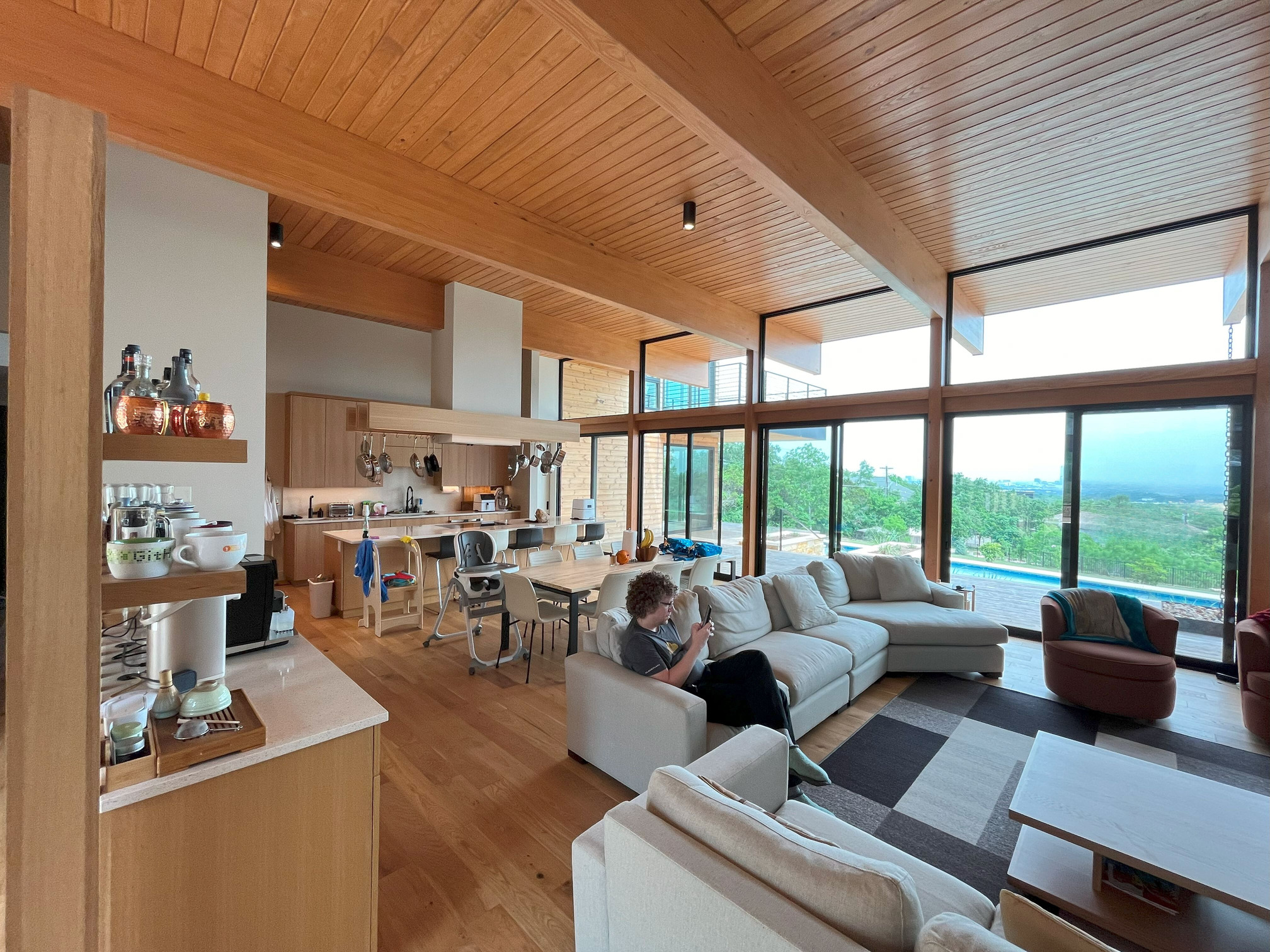 A photo depicts a large room, 40 feet long and 30 feet wide, which contains couches, a dining table, a full kitchen, and hardwood flooring. The ceiling is 14 feet high, held up by huge glulam beams. The far wall is mostly glass, composed of a row of sliding glass doors and transom windows above them. Beyond the glass is a distant view with lots of trees and hints of houses nestled between them. There is also a swimming pool, though it is barely visible. The kitchen features an island counter with an induction stovetop embedded within it. The vent hood above descends all the way from the ceiling. Wooden beams extend horizontally from the hood with pots and pans hanging from the beams. A series of bar stools on the opposite side allow people to sit at the island and chat with Jade while she cooks. The sink is along the back wall, making it difficult to keep Kenton company while he washes dishes.