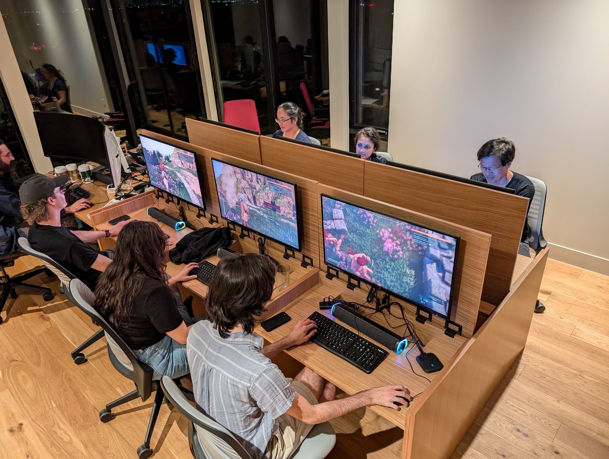 This photo depicts the same room as before, but now the conference table has transformed into six computer stations. The panels that formed the table surface before flip up, revealing a monitor mounted underneath. A second layed below them becomes the surface on which the keyboards sit. People are sitting at all the computers, playing video games. At the back of each station there is a slot where the keyboard and mouse are stored while not in use. Extending from the back side of the conference table is a normal-looking desk surface that does not fold up. Instead, a large curved monitor sits on top.