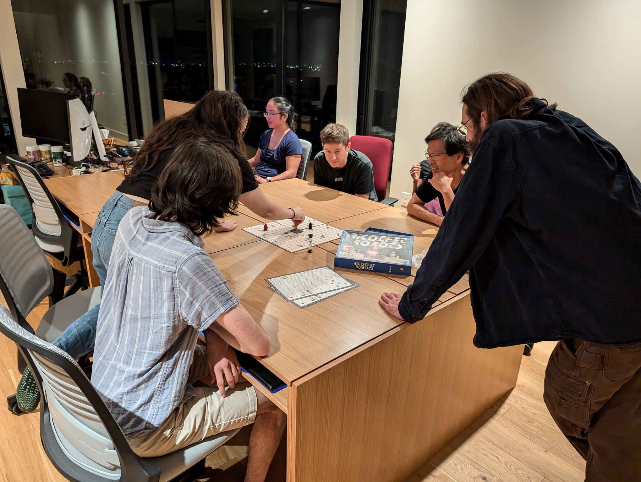 A photo depicts the office room, 16 feet wide and 20 feet long. Large windows on the far side show it is night time, with distant lights lining the horizon. In the middle of a room is a large wood conference table, aronud which several people are gathered, playing a board game. The table surface appears to be made up of several wood panels with cracks in between.