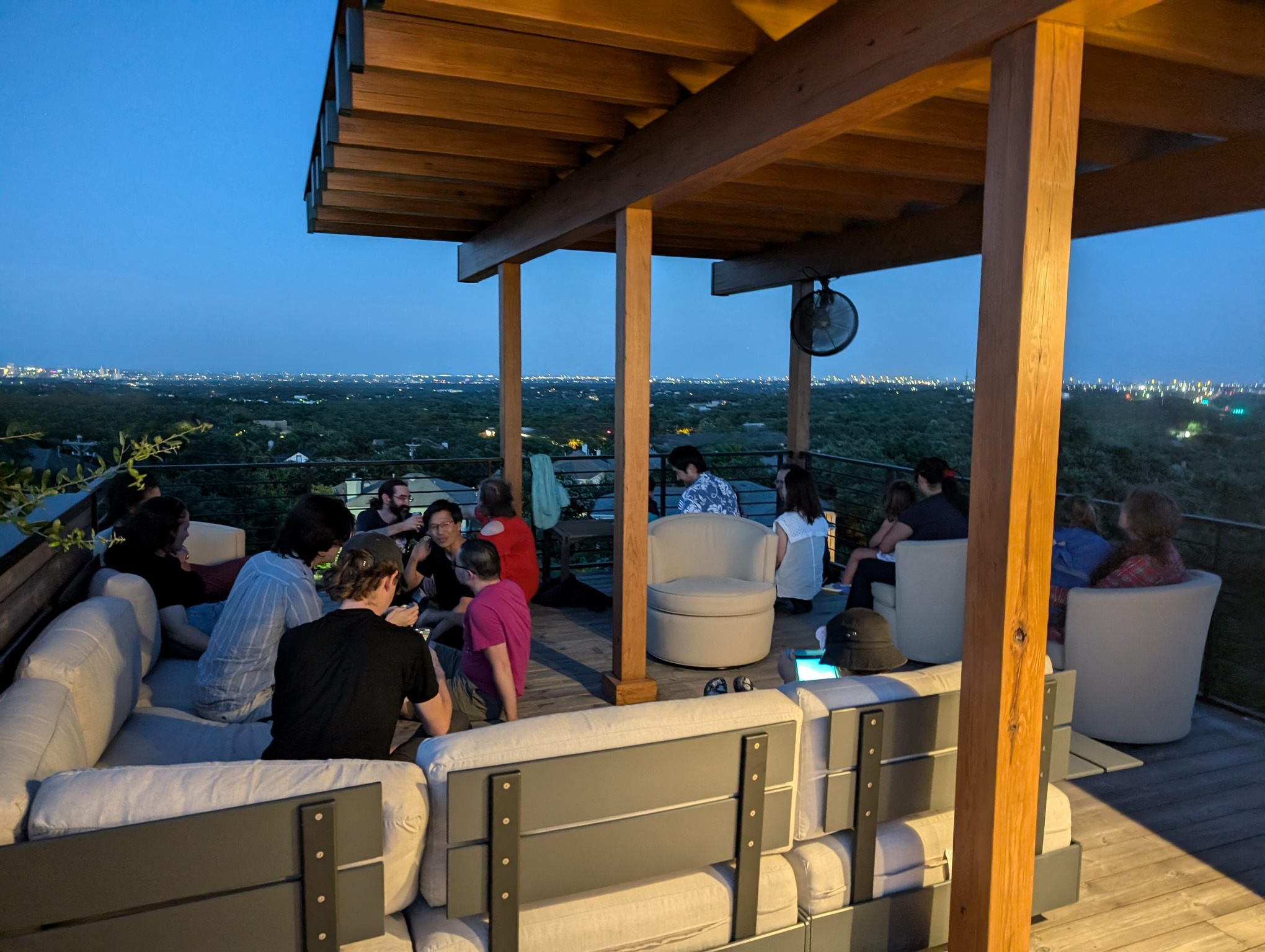 A photo depicts a roof deck with an epic view of trees, houses, and city lights in the distance. The deck hosts four swivel chairs and a large L-shaped sectional sofa. A covered trellis above the deck provides shade. Thirteen adults and four children are present, chatting and enjoying the view. Although not visible in the photo, the deck always experiences a pleasant breeze.